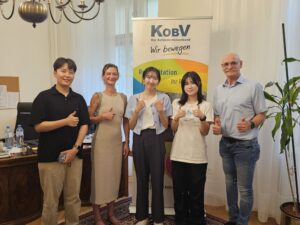 Gruppenfoto im Büro des KOBV Präsidenten. Links im Hintergrund der Schreibtisch aus dunklem Massivholz, dann die hohen Kastenfenster mit weißen Vorhängen. In der Mitte steht das KOBV Rollup mit dem Logo des KOBV Landesverbandes Wien, Niederösterreich und Burgenland. Davor von links nach rechts: Ein Vertreter der Pukyong National University mit schwarzem Haar, schwarzem Hemd und beiger Hose. Daneben die KOBV-Kommunikatorin mit braunem gewelltem Haar und grau-braunem Etuikleid. Neben ihr zwei Vertreterinnen der Pukyong National University. Die linkere trägt ihr schwarzes Haar zusammengebunden, hat ein hellblaues Hemd mit weißem T-Shirt und grauer Hose an. Die rechtere Dame trägt ihr schwarzes Haar offen über beide Schultern, und hat einen weißen Kimono und eine schwarze Hose an. Rechts außen steht KOBV Präsident Franz Groschan mit Brille, hellblauem Hemd und hellblauer Jeans. Alle 5 Personen halten ihre Daumen gehoben.