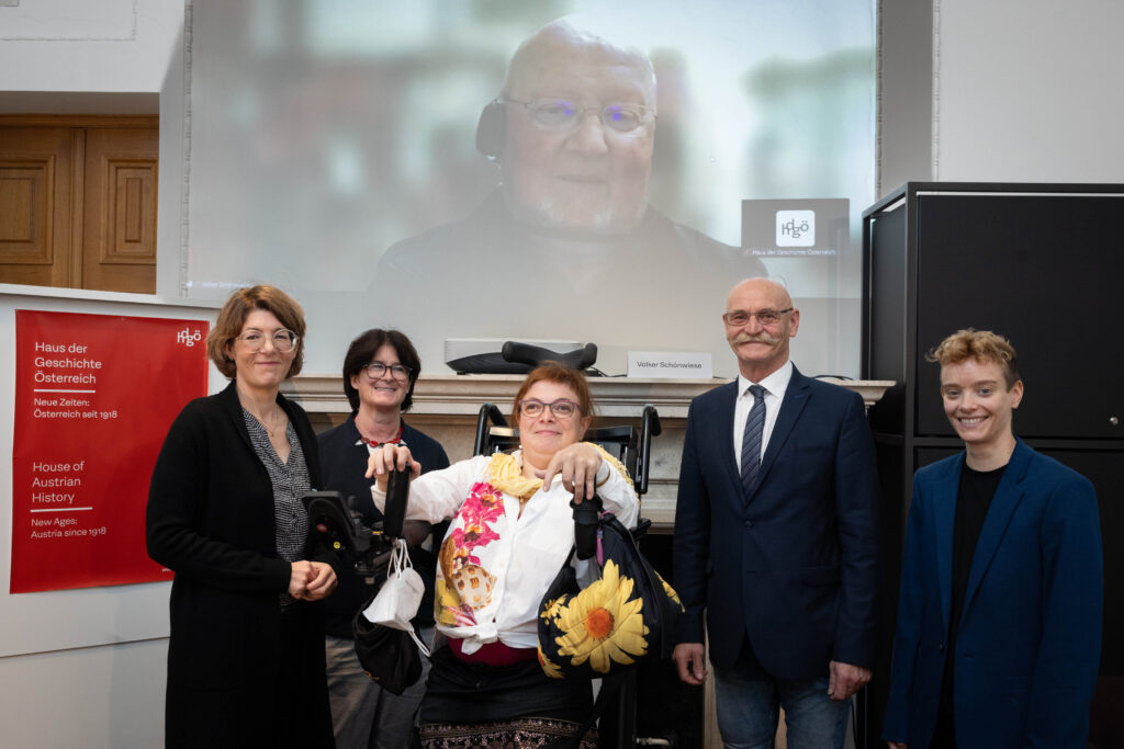 Die Fokusgruppe des Disability History Projects: Ganz links steht Frau Sommer mit schwarzer langer Weste, sie trägt eine schwarze Brille, daneben eine Dame mit schwarzen kurzen Haaren und Brille, Frau Margreiter im Rollstuhl sitzend und rechts steht Franz Groschan mit Glatze und Oberlippenbart, trägt schwarzes Sakko, Krawatte, Jeans und Brille, neben ihm Frau Tautter mit blonden kurzen Haaren und schwarzer Kleidung. Im Hintergrund ist Professor Schönwiese über eine Leinwand online zugeschalten. Er hat eine Glatze und trägt Brille.