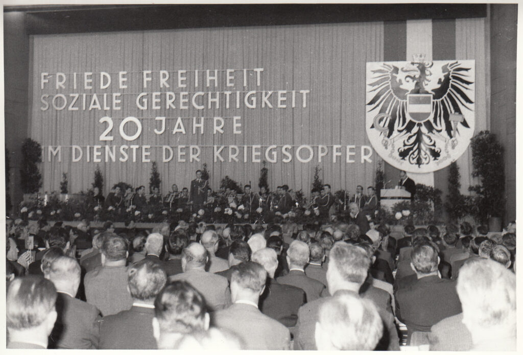 Ein schwarz-weiß Foto eines Delegiertentages. Musikanten in Uniform sind am mit Sträuchern geschmückten Podium, ein Herr mit Brille steht am Rednerpult, an der Wand über dem Podium ist in großer Schrift zu lesen: Friede Freiheit soziale Gerechtigkeit 20 Jahre im Dienste der Kriegsopfer. Daneben hängt ein riesiges Schild des österreichischen Doppeladlers auf österreichischer Flagge. Im Vordergrund das Publikum von hinten, allesamt Herren.