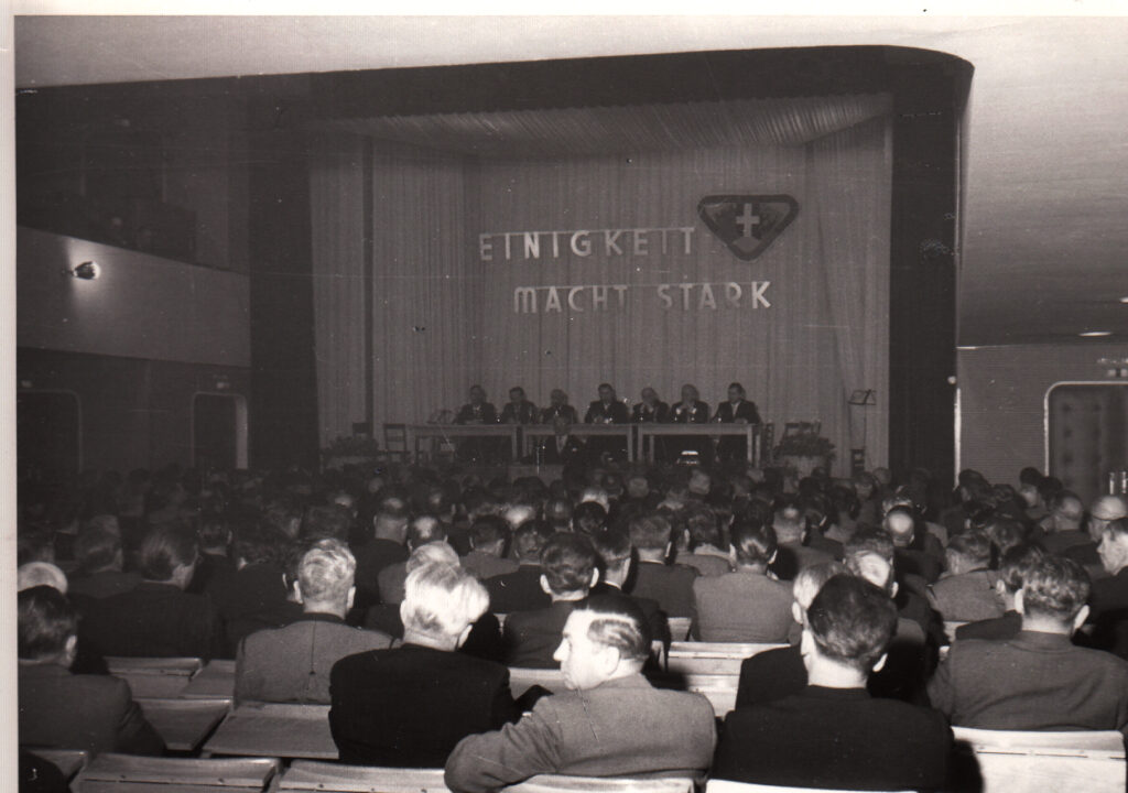 Ein altes schwarz-weiß Foto vom 7. Delegiertentag des damaligen KOV mit 7 Männern am Podium und Publikum in den Bankreihen, das von hinten abgebildet ist. Über dem Rednertisch ist in großen funkelnder Schrift zu lesen: Einigkeit macht stark.