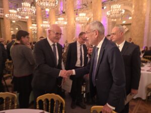 KOBV-Präsident Franz Groschan und Bundespräsident Alexander van der Bellen schütteln einander freundlich lächelnd die Hände. Sie stehen im prunkvollen Festsaal im Historismus-Stil der Hofburg Wien.