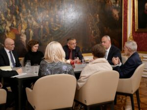 Am großen ovalen Holztisch der Präsidentschaftskanzlei vor dem großem Wandgemälde mit sehr vielen Personen darauf, sitzen Franz Groschan, Christine Schneyder, Bundespräsident van der Bellen und mit dem Rücken zur Kamera First Lady Doris Schmidauer und Magistra Christine Steger links neben ihr.